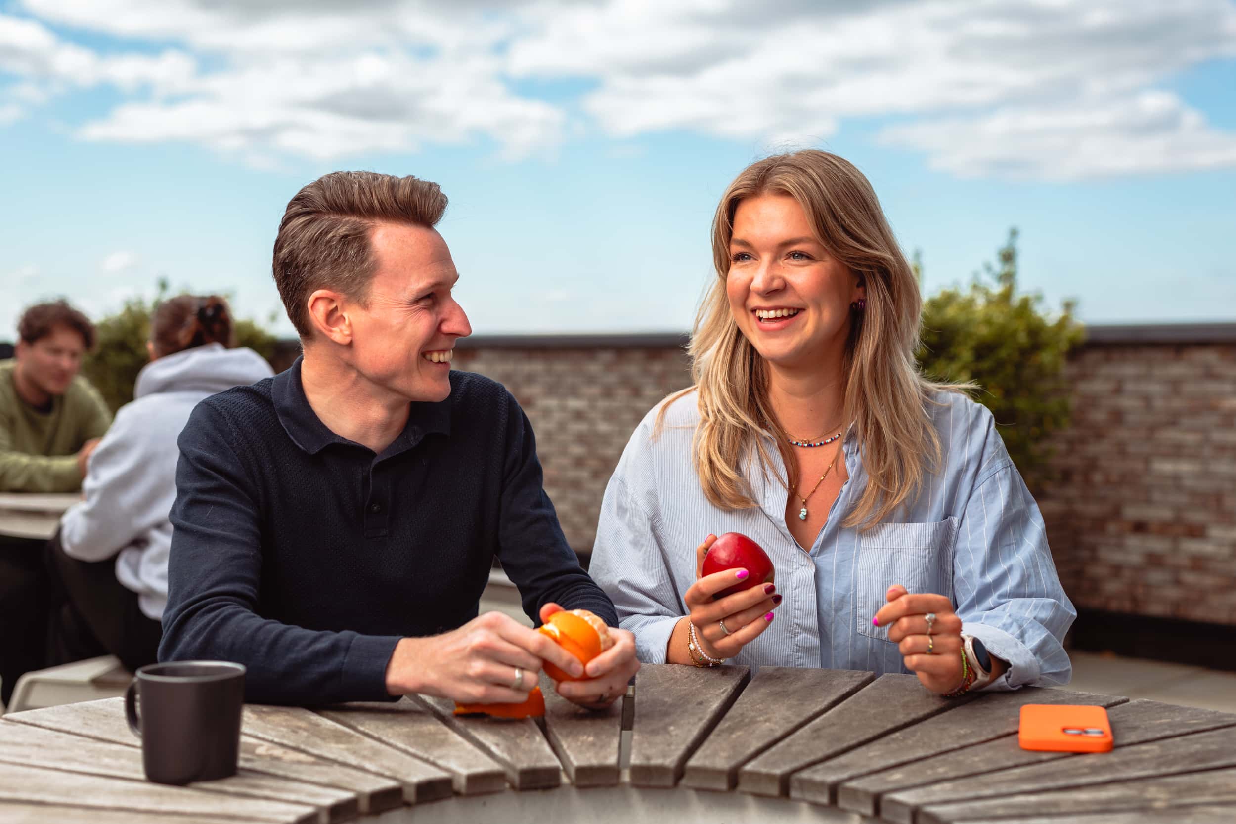 Twee collega's lachen naar elkaar en zitten op een dakterras met een stuk fruit in hun hand.