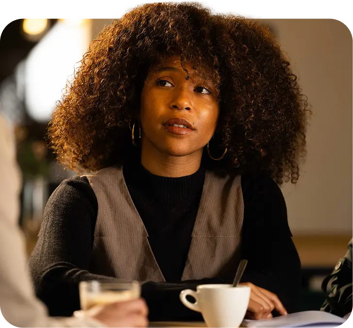 Een vrouw met krullend haar zit aan tafel, gekleed in een zwarte top met een beige vest. Voor haar staat een kop koffie. Ze is in gesprek met anderen, die deels zichtbaar zijn.