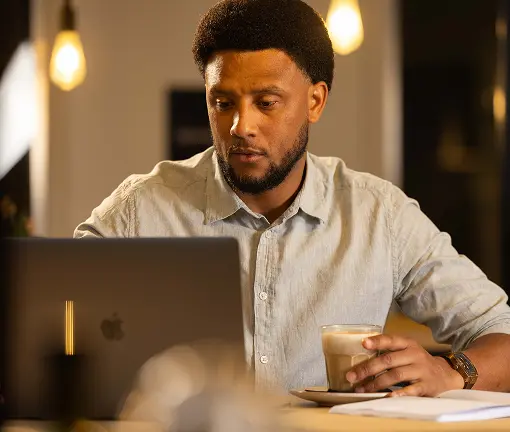 Een man zit aan een tafel in een sfeervol verlichte ruimte, kijkt geconcentreerd naar zijn laptop en houdt een glas koffie vast, met een open notitieboek voor zich.