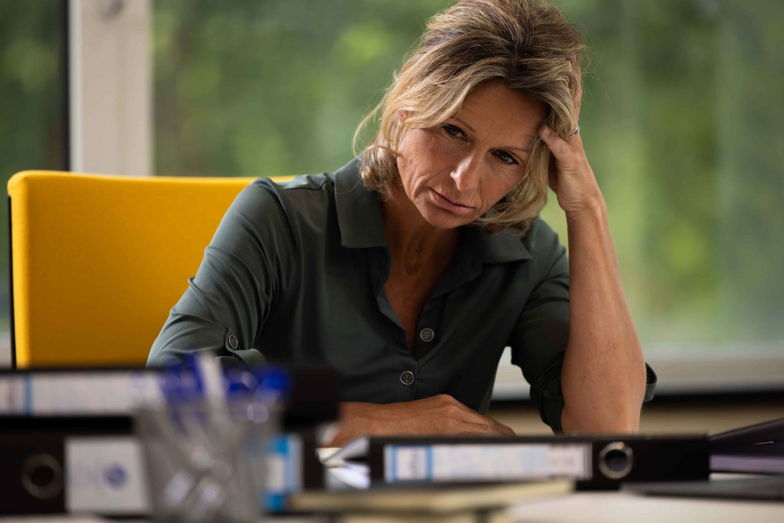 Een dame zit aan een bureau met één hand op het bureau en de andere hand tegen het hoofd. Ze heeft stress. Op de achtergrond is een raam met uitzicht op groen zichtbaar. Op het bureau liggen ordners en kantoorspullen.