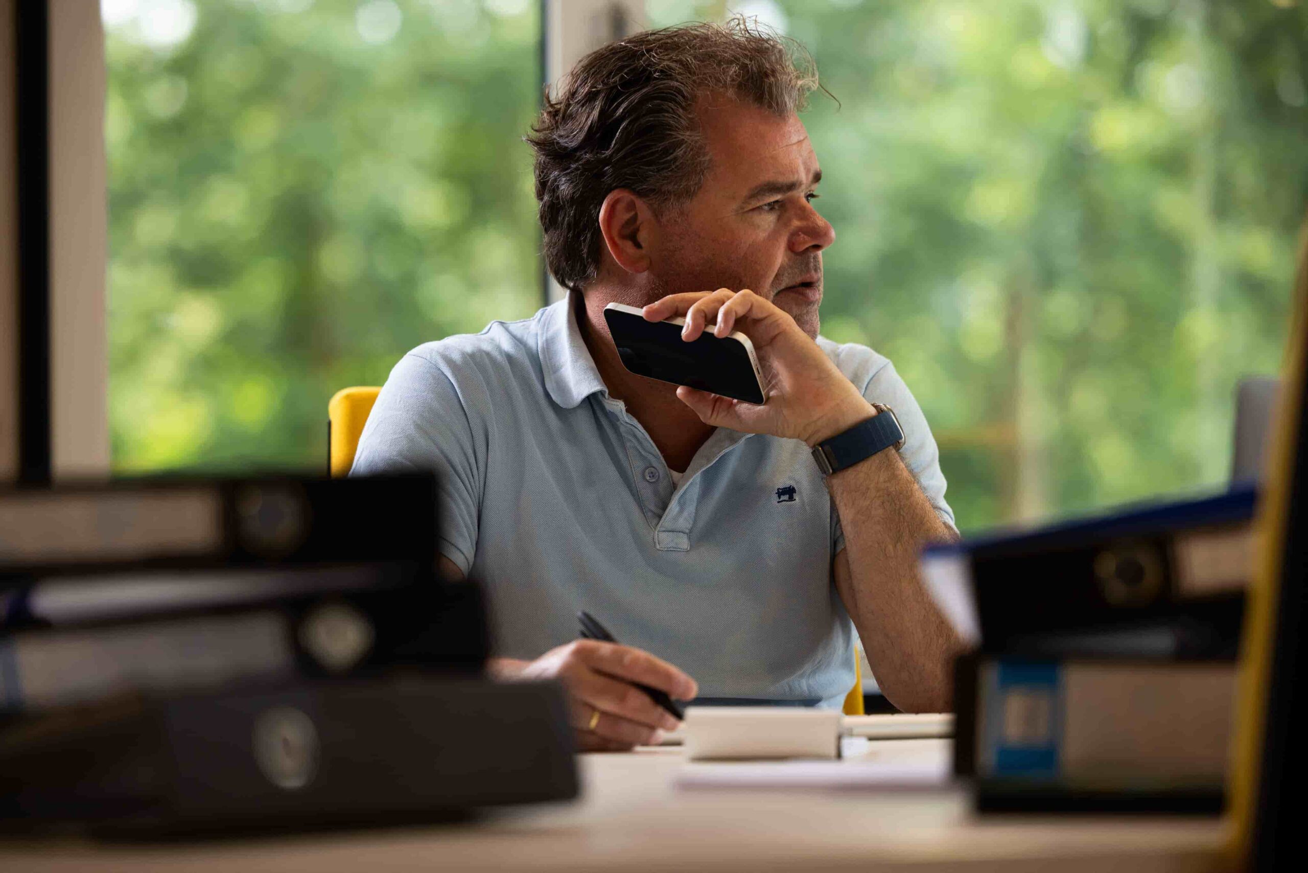 Medewerker is vermoeid en gestrest met zijn telefoon in de hand. Er ligt een hoop werk voor zich op het bureau.