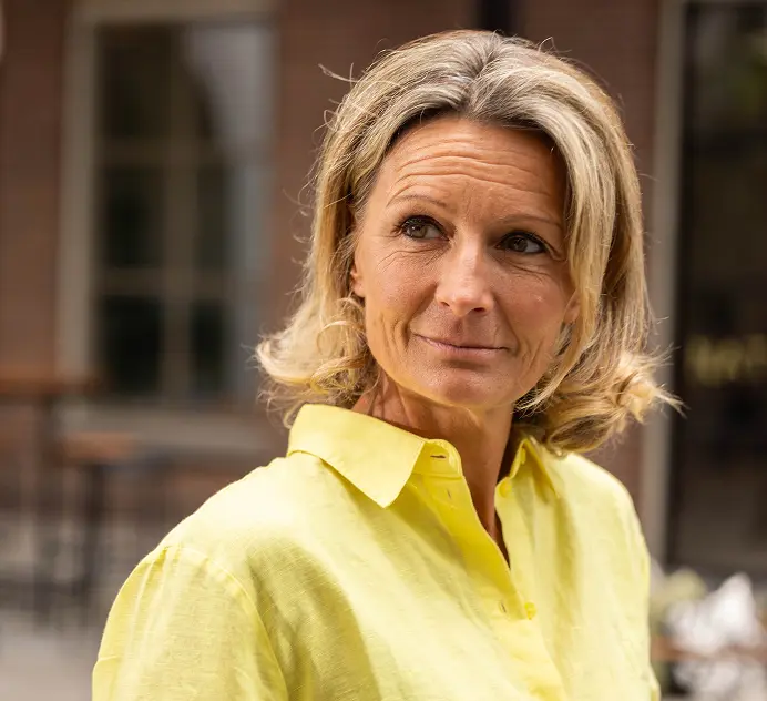 Een dame in een geel shirt staat buiten en houdt een beker vast. Op de achtergrond zijn vage contouren van tuinmeubilair en een gebouw met grote ramen zichtbaar.
