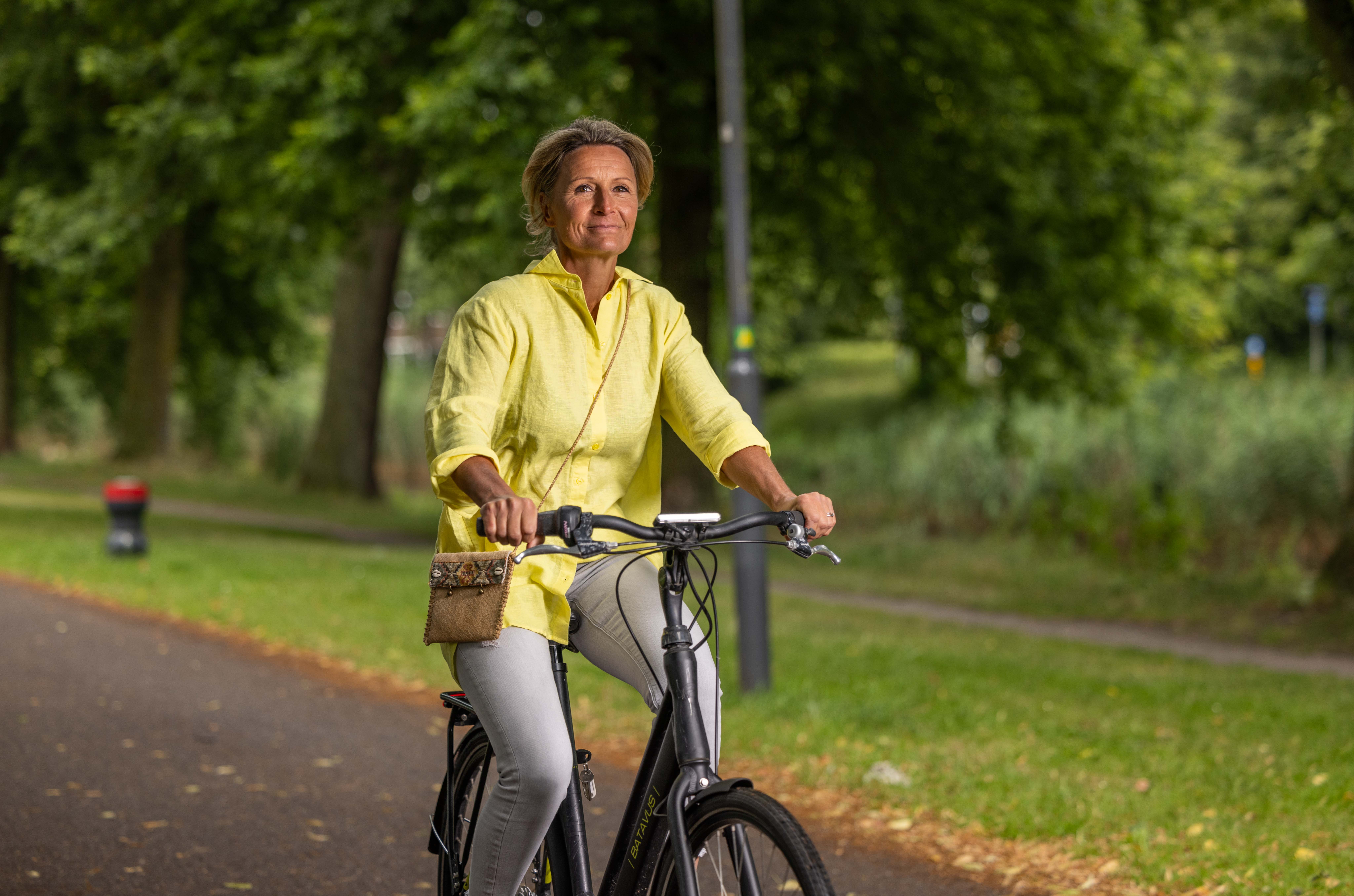 Medewerkster komt met de fiets naar het werk en fietst door het park. Ze heeft haar tas bij zich en kijkt vrolijk