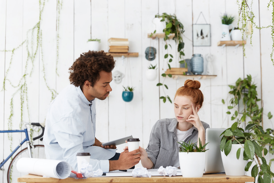 Twee personen in een werkruimte met planten en planken op de achtergrond. Eén persoon zit aan een bureau met een laptop, terwijl de ander staat en een beker aanreikt. Op het bureau liggen opgerolde papieren, verfrommelde papiertjes en koffiebekers.
