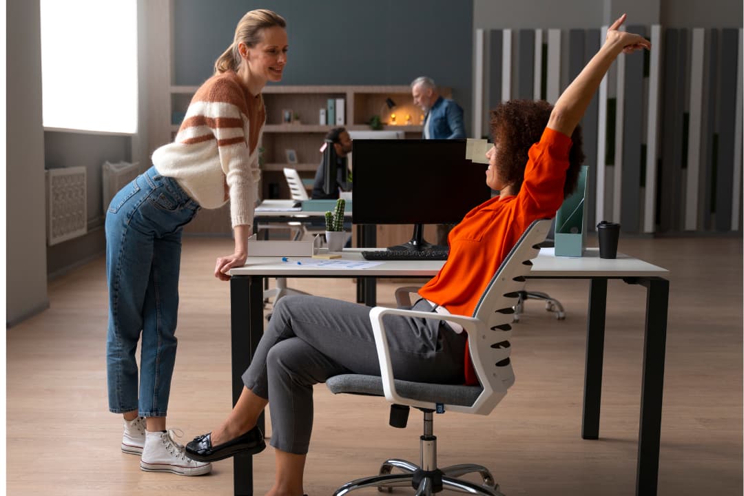 In een kantooromgeving staat één persoon leunend op een bureau, terwijl een ander persoon zittend in een stoel een arm omhoog strekt. Op de achtergrond zijn bureaus, computers en kantoorspullen zichtbaar.