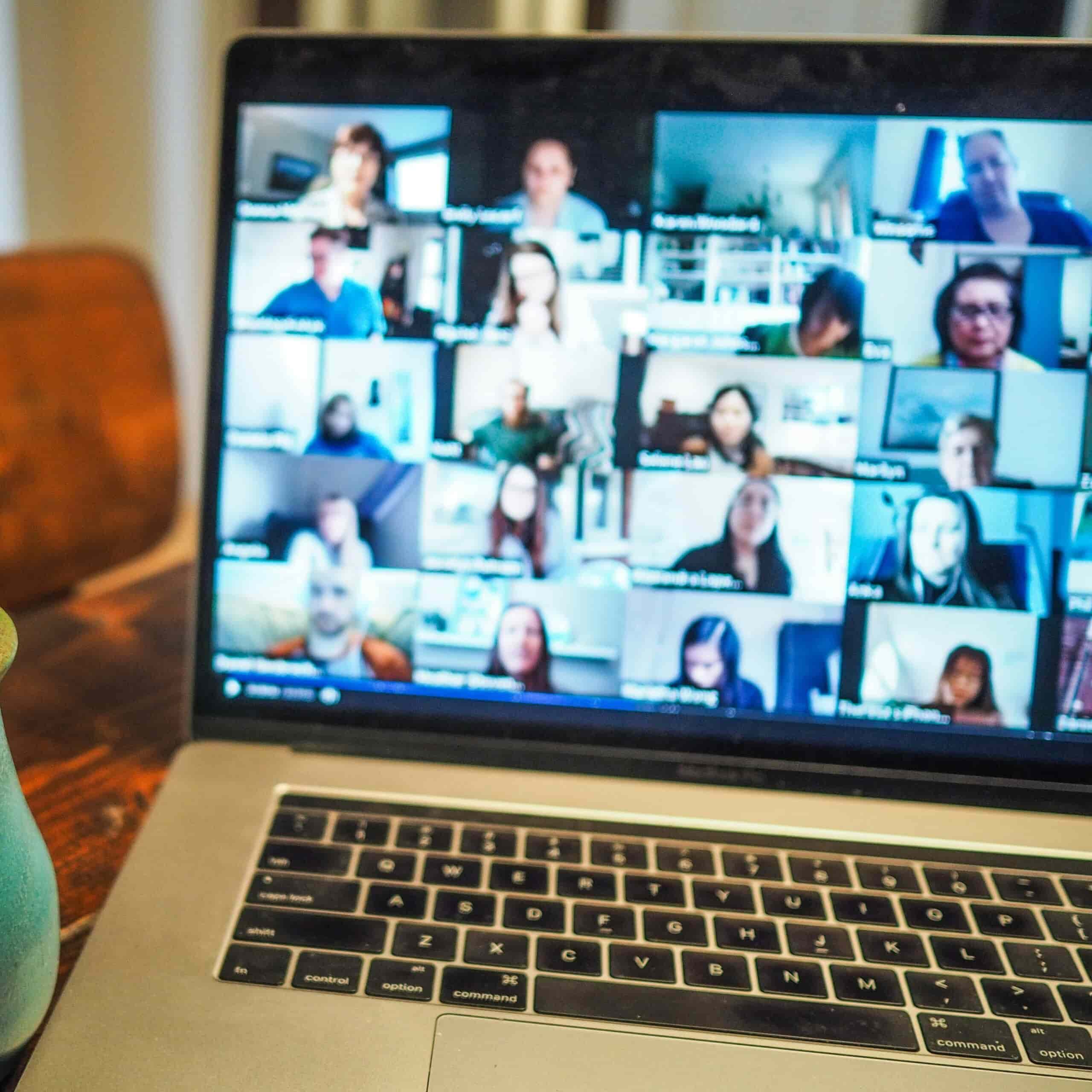 Een laptopscherm toont een videogesprek met meerdere deelnemers, elk zichtbaar in een eigen venster in een rasterindeling. De gezichten zijn vervaagd voor privacy.