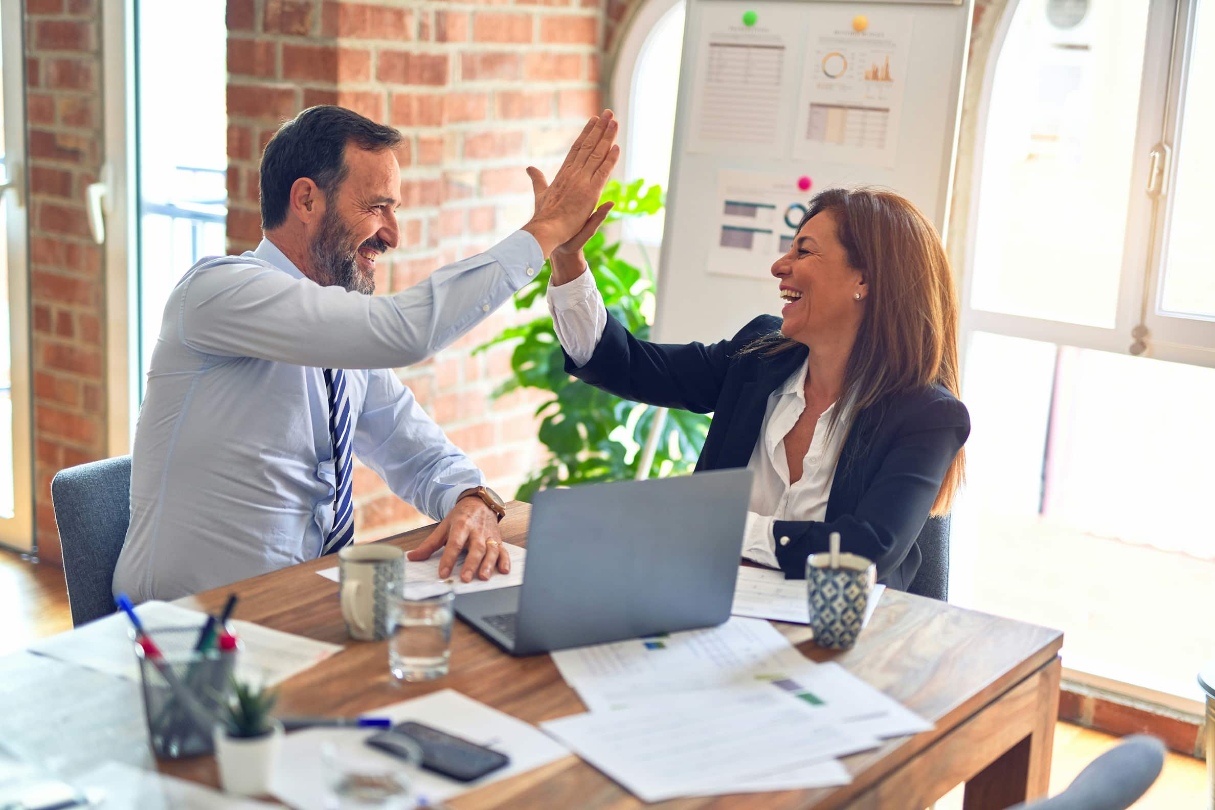 Twee personen zitten aan een houten tafel in een kantooromgeving en geven elkaar een high-five. Op de tafel liggen papieren, een laptop, mokken en een plant. Op de achtergrond is een whiteboard te zien met grafieken en diagrammen.