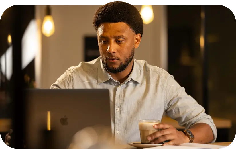 Een man zit aan een tafel in een sfeervol verlichte ruimte, kijkt geconcentreerd naar zijn laptop en houdt een glas koffie vast, met een open notitieboek voor zich.