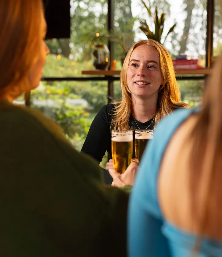 Drie collega's zitten aan een tafel in bar. De dame in het midden houdt een glas 0.0 bier vast. Op de achtergrond zijn grote ramen met uitzicht op groen en decoratieve elementen zoals planten en hanglampen te zien.