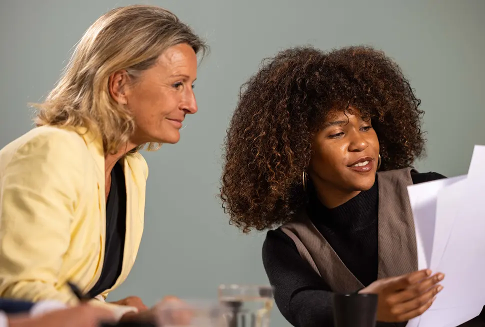 Twee personen zitten aan een tafel en bekijken samen documenten. De persoon links draagt een gele blazer, terwijl de persoon rechts met krullend haar de papieren vasthoudt.